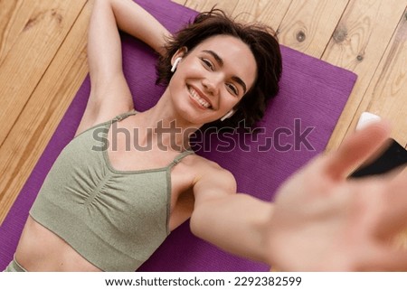 young pretty woman doing yoga at home wearing stylish sport outfit watching exercises on phone, meditation on mat, listening to music in earpods