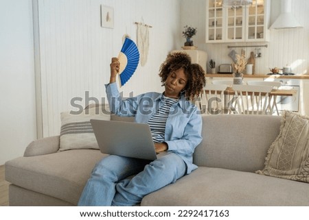 Working from home in heatwave. Exhausted overheated African American girl freelancer waving hand fan working online on laptop, unhappy black woman trying to cool down at home office during summer heat Royalty-Free Stock Photo #2292417163