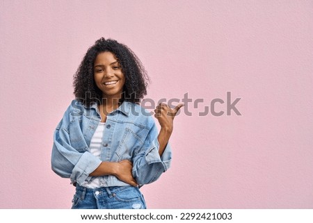 Portrait of teenage african american hipster girl in denim clothes and with curly afro hairstyle stand and point on copy space for advertising, looking at camera and smiling on soft pink background. Royalty-Free Stock Photo #2292421003