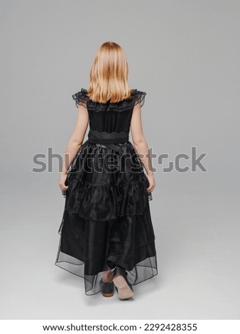 rear view. cute blonde teen girl in a black long curvy dress on a gray background. the concept of elegant clothes for children. cosplay.