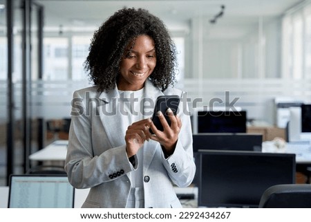 Young smiling successful African American business woman, beautiful female entrepreneur businesswoman using smartphone, cellphone application, online communication, standing in modern office indoors.