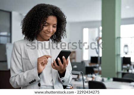 Young smiling successful African American business woman, beautiful female entrepreneur businesswoman using smartphone, cellphone application, online communication, standing in modern office indoors.