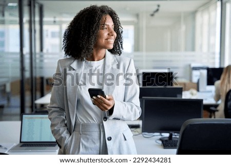 Young smiling successful African American business woman, dreaming female entrepreneur businesswoman using smartphone, cellphone application, online communication, standing in modern office indoors.