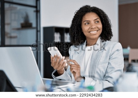 Young smiling successful African American business woman, beautiful female entrepreneur businesswoman using smartphone, cellphone application, online communication, sitting in modern office indoors.
