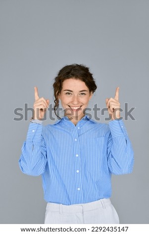 Happy young professional business woman wearing blue shirt looking at camera pointing finger up at copy space showing upward presenting advertising standing isolated at gray background, vertical.