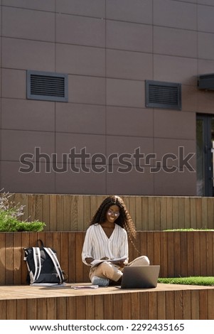 Black young woman model, pretty curly African female university student elearning using laptop computer writing notes studying outside campus on sunny day, remote learning, vertical shot.