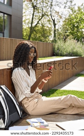 Young smiling happy black African woman university student model holding cell phone using mobile apps, looking at smartphone, typing on cellphone, chatting sitting outdoors campus, vertical shot.
