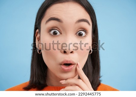 Close up portrait of girl looks with surprised, amazed eyes, says wow, impressed by smth, stands over blue background. Royalty-Free Stock Photo #2292445775