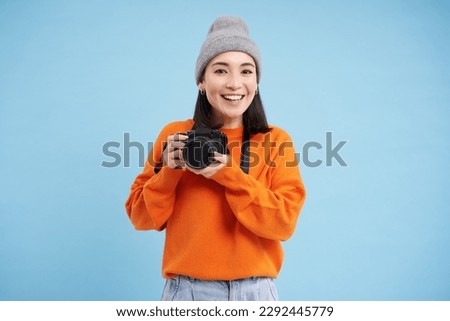 Stylish asian girl with digital camera, taking pictures. Woman photographer smiling, standing over blue background.