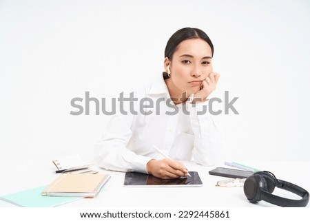 Tired and bored korean woman in office, employee sits with digital tablet, listens music in earphones and looks tired, white background.