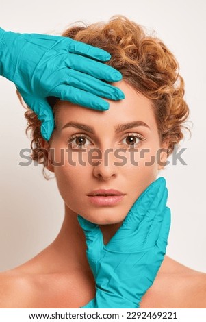 Close-up portrait of woman's face, young brown-eyed curly woman looks at camera, hands in blue medical gloves touch her face forehead and chin, health care concept, copy space, high quality photo