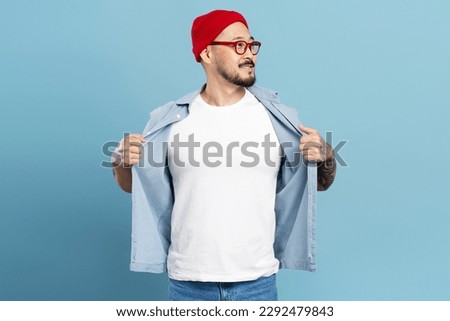 Smiling confident asian man, stylish hipster wearing red hat, eyeglasses, showing white t shirt with mockup. Korean model posing for pictures, isolated on blue background. Store, shopping concept