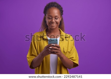 Young positive beautiful African American woman in casual clothes typing text on mobile phone using dating apps or instant messengers to find boyfriend stands on isolated lilac background Royalty-Free Stock Photo #2292487141