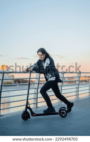 A young man with long hair of an informal appearance rides along the embankment on an electric scooter.