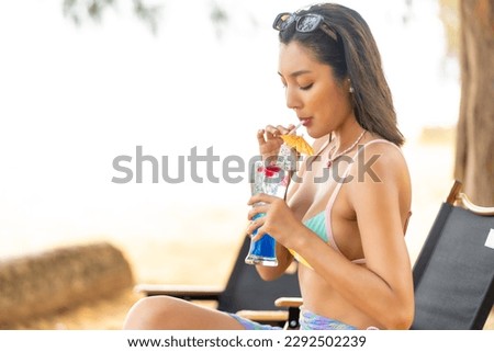 Happy Asian woman travel nature ocean on summer holiday vacation. Beautiful girl in swimwear bikini enjoy outdoor lifestyle drinking cocktail at beach cafe at tropical island beach in sunny day.
