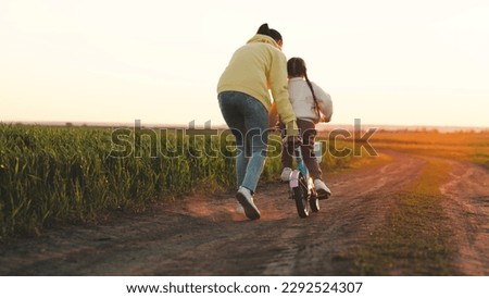 mother rides child bicycle sunset. happy family park. Mom teaches her daughter ride bike. concept happy family summer park. fun vacation. teamwork. mother runs green grass rolling child bicycle. Royalty-Free Stock Photo #2292524307