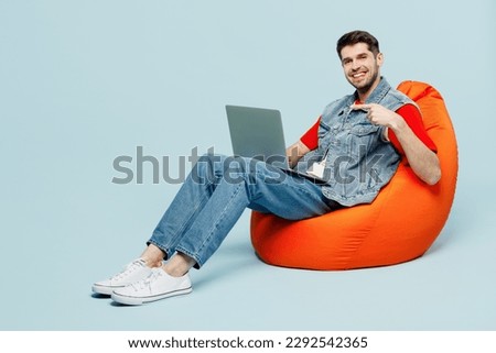Full body fun young IT man he wear denim vest red t-shirt casual clothes sit in bag chair hold use work poiint finger on laptop pc computer isolated on plain pastel light blue cyan background studio