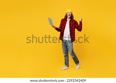 Full body young smiling happy caucasian IT man wearing red checkered shirt white t-shirt hat hold use work on laptop pc computer do winner gesture isolated on plain yellow background studio portrait