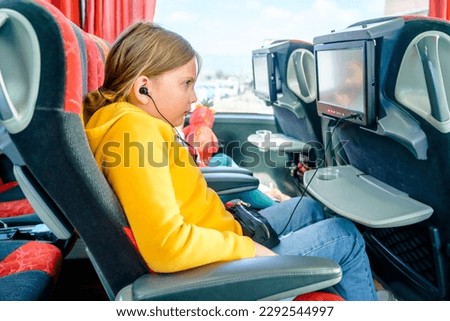 Bus road traveling. Girl kid rides on large comfortable sightseeing excursion bus. Watching movie cartoon on screen in chair. Entertainment media system. Sittting, listening to music in headphones.