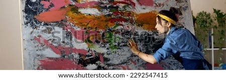 Creative young female painter working on a large modern abstract oil painting, applying paint on canvas with fingers in living room at home studio workshop