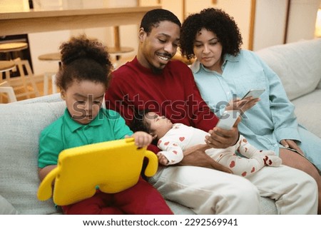 Happy loving multicultural family black father, hispanic mother, two little kids sitting on couch at home, using gadgets, man and woman holding smartphones, toddler watching cartoon on digital tablet