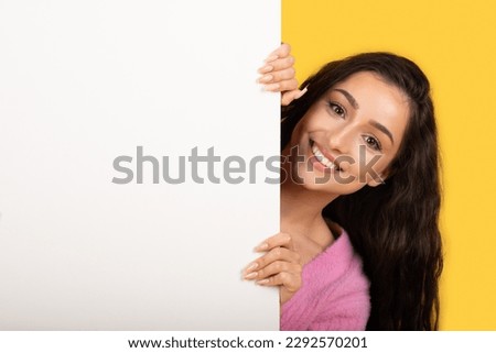 Smiling young arab lady peeks out from behind big banner with empty space for ad, text and offer isolated on yellow background, studio, close up. Recommendation, sale, presentation