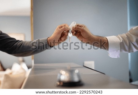 Unrecognizable Hotel Receptionist Giving Key To Male Guest At Reception In Lobby, Closeup Of Hands. Tourist Checking In At Accomodation, Taking Room Keys From Hostel Worker. Cropped Shot