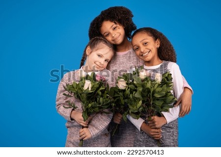 Cheerful teen african american and caucasian girls with bouquets of flowers hugs, enjoy spring isolated on blue studio background. Fun, holiday celebration, childhood and