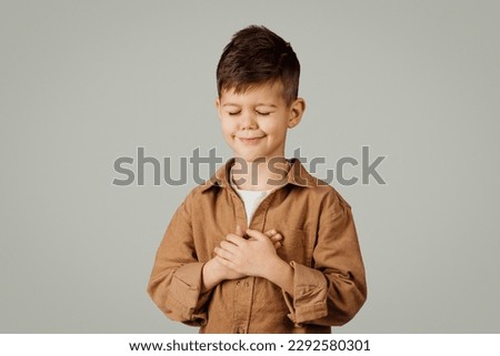 Positive cute little european child 6 years old in casual with closed eyes puts hands to chest, prays, makes wish isolated on gray background. Education, gratitude, childhood, study and emotions