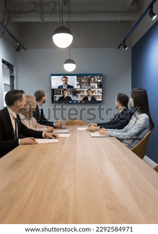 Team of businesspeople meeting at table in small conference room, discussing work project with remote diverse colleagues on group video call, turning heads to screen with facial head shots Royalty-Free Stock Photo #2292584971