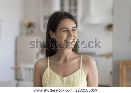 Cheerful dreamy late teenage girl indoor candid portrait. Beautiful young Latin woman with healthy white teeth smiling, looking at window away, dreaming, thinking on achievement, success