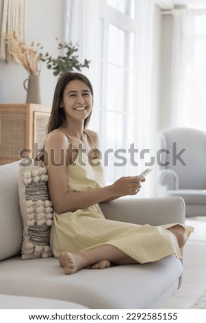 Happy beautiful adult Hispanic girl holding mobile phone, sitting on home sofa, looking at camera with toothy smile, laughing, using Internet connection technology. Vertical portrait