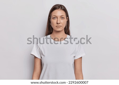 Waist up shot of nice looking woman has dark hair dressed in basic t shirt keeps arms down looks directly at camera isolated on white background. Brunette female model poses in studio for making photo