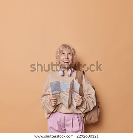 Travel concept. Positive blonde young European woman holds map and dreams about journey concentrated above happily dressed casually poses with rucksack isolated over brown background copy space
