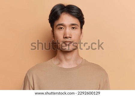 Portrait of serious dark haired adult Asian man with neutral facial expression focused at camera dressed in casual jumper isolated over brown background. Handsome Japanese guy poses in studio Royalty-Free Stock Photo #2292600129