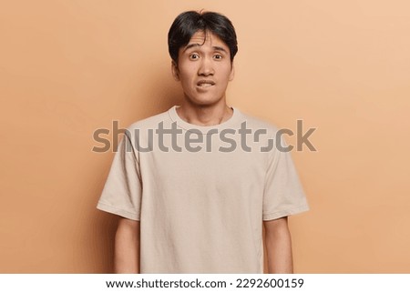 Waist up shot of worried Japanese man bites lips looks anxious and surprisingly at camera doesnt know way out from difficult situation dressed in casual t shirt isolated over brown background Royalty-Free Stock Photo #2292600159