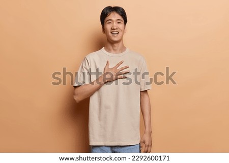 Photo of cheerful young Asian man presses hand to chest expresses gratitude reacts to something cute expresses kindness and sympathy dressed in casual t shirt poses against brown background.