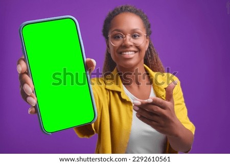 Young excited beautiful African American woman holds out mobile phone to camera pointing at green screen to advertise your application or web service posing on isolated purple background