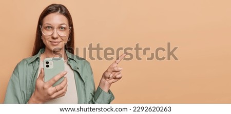 Indoor shot of pretty dark haired young woman holds mobile phone asks to check this out points index finger uses mobile phone wears big spectacles and green shirt isolated over brown background