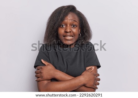 Indoor shot of dark skinned woman trembles from cold hugs herself feels cold as wears only t shirt feels scared notices something terrifying isolated on white background. Oh its very freezing outside