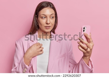 Horizontal shot of serious busy brunette female office worker chats on web uses mobile phone for video conference has job interview distantly dressed in formal outfit poses against pink background