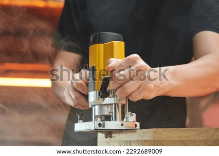 The hands of a Caucasian worker lead an electric milling cutter along the edge of a wooden blank. the milling machine processes the parts and raises the chips and smoke into the air Royalty-Free Stock Photo #2292689009