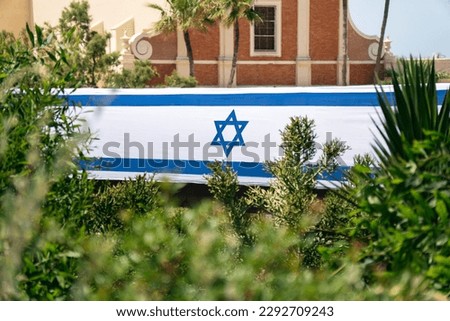View of the Israeli flag through the leaves of plants in the old city of Jaffa in honor of Independence Day with the wall of the ancient church in the background