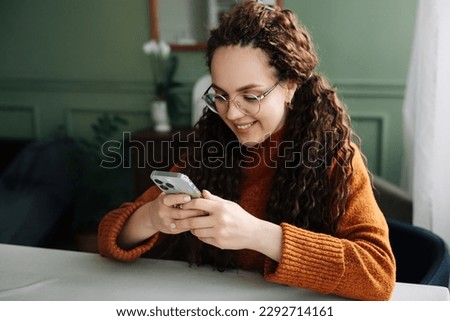 Young smiling woman wearing glasses holding a smartphone using cellphone modern technology, looking at mobile, checking cell phone apps, texting and browsing the internet for shopping sitting at home Royalty-Free Stock Photo #2292714161