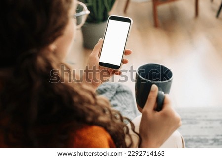 Over-shoulder view of woman holding coffee and smartphone with white mock-up cellular screen in hands using mobile apps on a cell phone. Cellphone display mock-up for advertising concept. Royalty-Free Stock Photo #2292714163