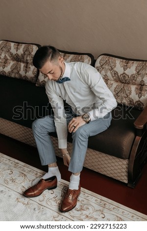 portrait. A man in a white shirt and gray pants is sitting on the sofa and putting on his elegant shoes. A stylish watch. Men's style. Fashion. Business