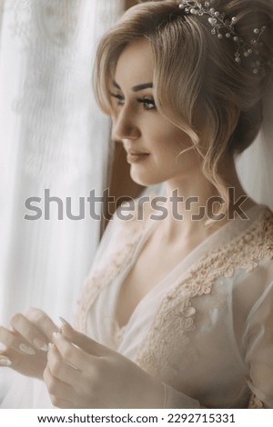 A bride with an elegant hairdo and tiara, in a robe, stands holding her ring and looking at it. Beautiful makeup. Wedding photo. Blonde