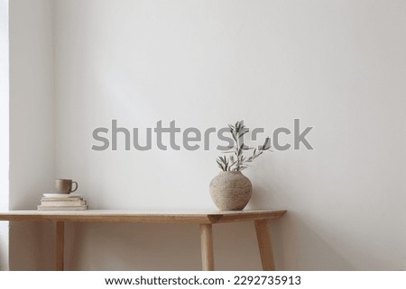 Elegant Mediterranean home design. Textured vase with olive tree branches, cup of coffee. Books on wooden table. Living room still life. Empty wall copy space. Modern interior, no people. Home office.