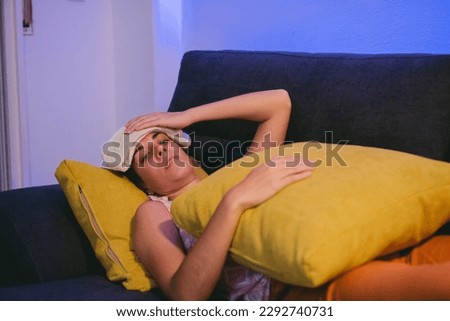 Sick young Latin woman lying on a sofa with a towel on her head to bring down her fever while hugging a cushion. Royalty-Free Stock Photo #2292740731
