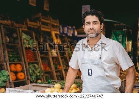 Middle-aged Latin man in an apron looking at the camera in the greengrocer's shop where he works. Copy space. Royalty-Free Stock Photo #2292740747